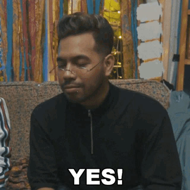 a man wearing glasses is sitting on a couch and says " yes "