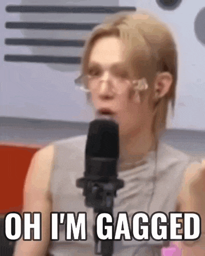 a man wearing glasses is talking into a microphone and says `` oh i 'm gagged '' .