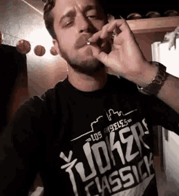 a man wearing a black t-shirt that says joker classick is smoking a cigarette