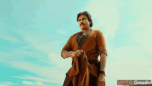 a man in a brown shirt stands in front of a blue sky with the words vara gaadu written below him
