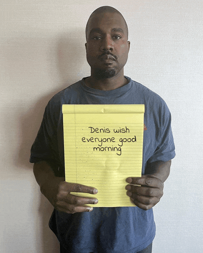 a man in a blue shirt holds up a yellow notepad that says denis wish everyone good morning