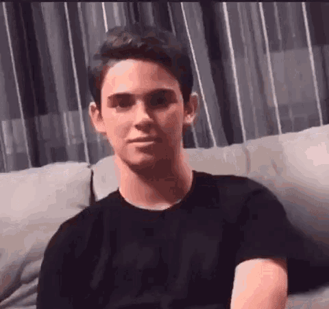a young man in a black shirt is sitting on a couch and making a funny face .