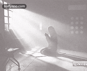 a woman is kneeling down in front of a window in a dark room praying .