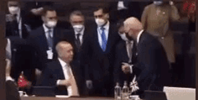 a group of men in suits and ties are standing around a table . one of the men is wearing a mask .