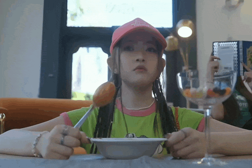 a girl wearing a pink hat and a green shirt is eating from a bowl with a fork