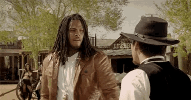 a man with dreadlocks and a cowboy hat is talking to another man in a western town .