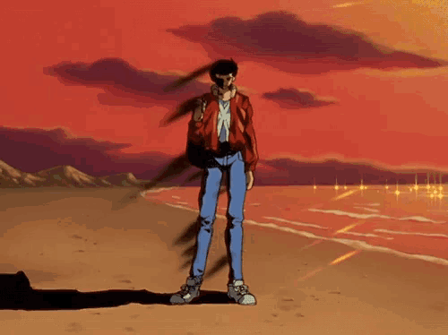 a man in a red jacket and blue jeans stands on a beach at sunset