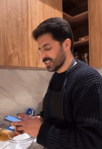 a man with a beard wearing an apron is using a cell phone in a kitchen .