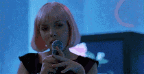 a woman with pink hair is singing into a microphone in front of a television .