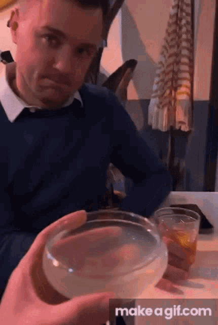 a man in a blue sweater is sitting at a table holding a glass of liquid in front of him