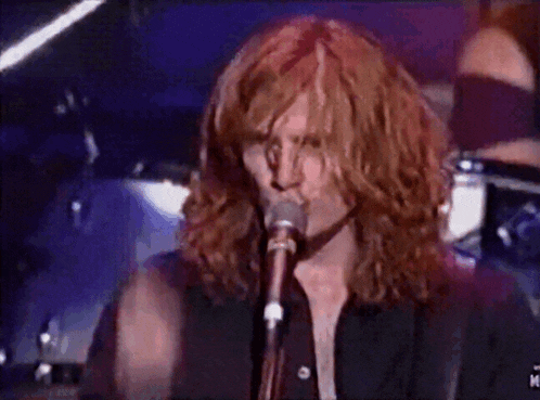 a man with long red hair singing into a microphone with the letter m on the bottom right