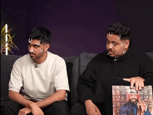 two men are sitting on a couch and one is holding a picture of a man with a beard