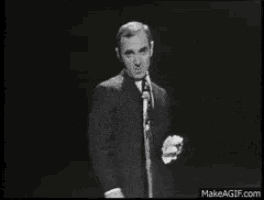 a man in a suit and tie is standing in front of a microphone and pointing at the camera .