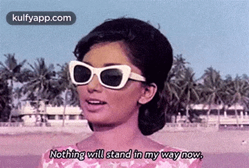 a woman wearing sunglasses is standing on a beach and saying nothing will stand in my way now .