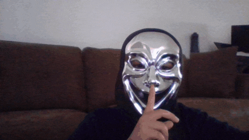 a man wearing a silver anonymous mask holds his finger to his lips
