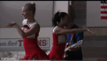 two cheerleaders are dancing in a hallway with a man holding a guitar .