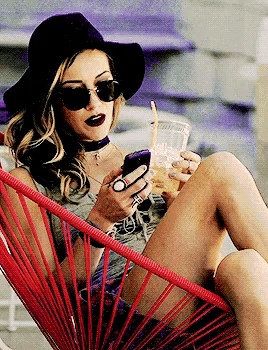 a woman wearing a hat and sunglasses is sitting in a red chair holding a drink and looking at her phone