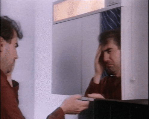 a man looks at his reflection in a mirror while holding his head