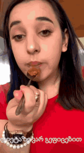 a woman in a red shirt is eating something with a spoon and a watch on her wrist