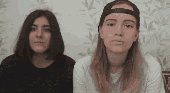 two young women are standing next to each other and looking at the camera . one of the women is wearing a baseball cap .