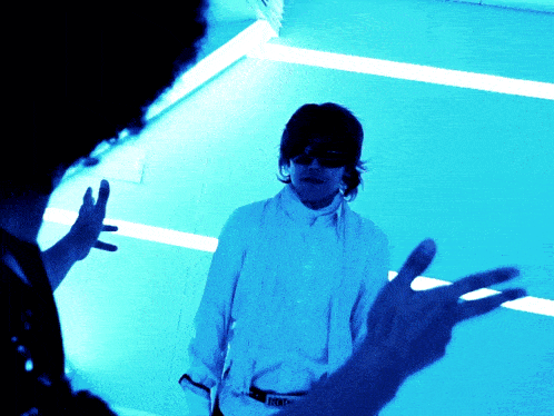 a man wearing sunglasses and a white shirt talks to another man in a blue room