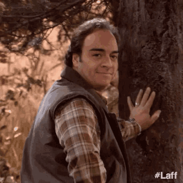 a man in a plaid shirt and vest is touching a tree .