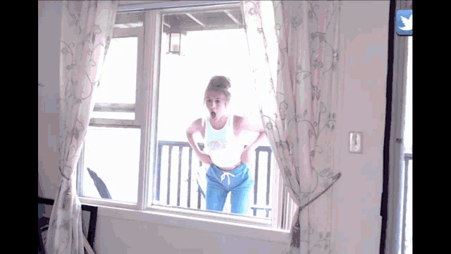 a woman in a white tank top is standing in front of a window looking out