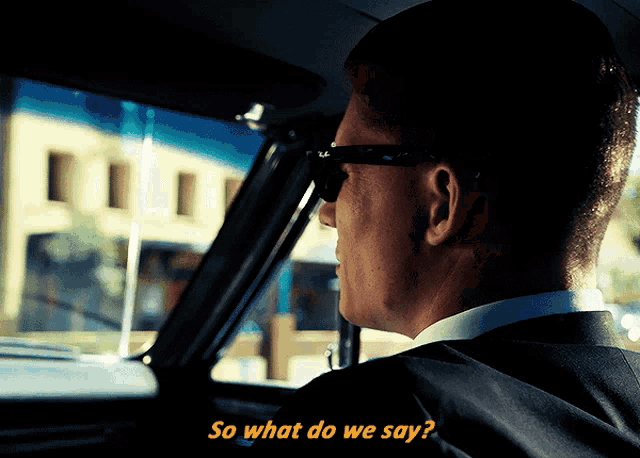 a man in a suit and sunglasses is driving a car and says so what do we say