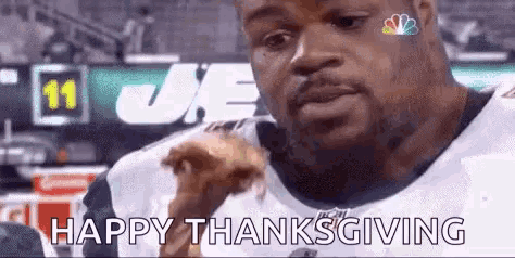a man in a football uniform is eating a chicken wing and saying happy thanksgiving .