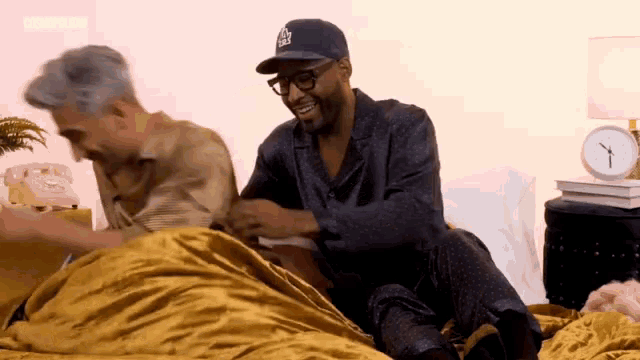 a man wearing a la dodgers hat sits on a bed with another man