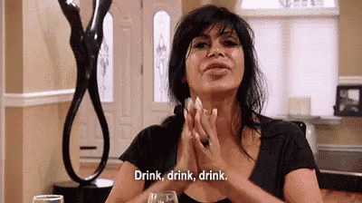 a woman is sitting at a table with a glass of wine and saying `` drink , drink , drink '' .