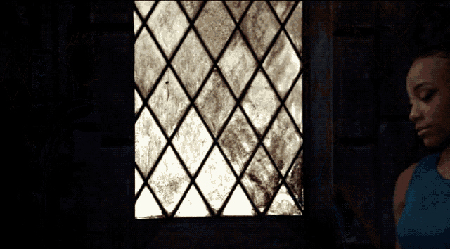 a woman in a blue tank top looks out a dirty stained glass window