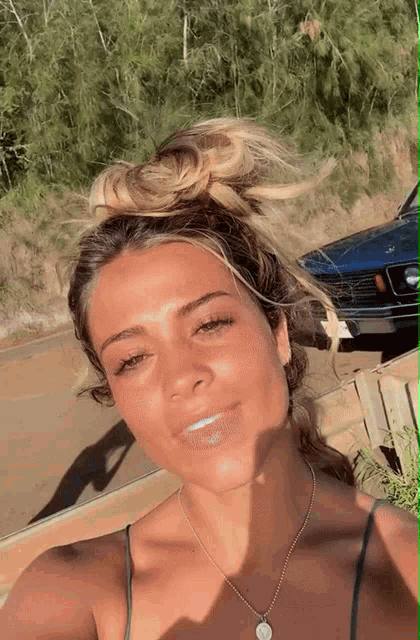 a woman wearing a necklace and a bun takes a selfie in front of a blue car