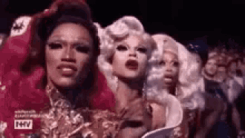 a group of drag queens are standing next to each other in front of a crowd of people .