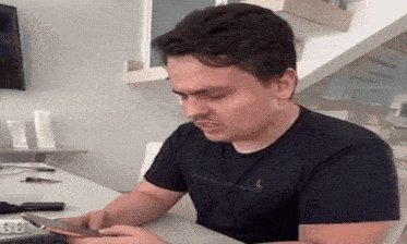 a man in a black shirt is sitting at a table looking at his cell phone .