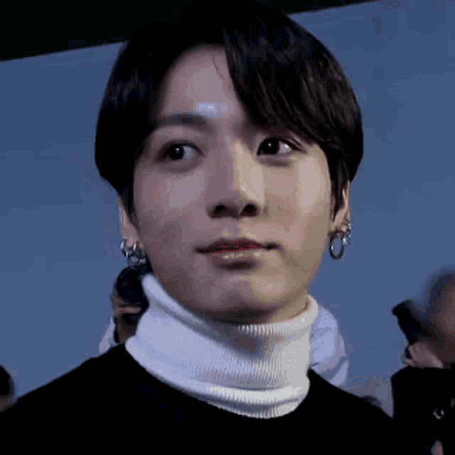 a close up of a man wearing a white turtleneck and earrings
