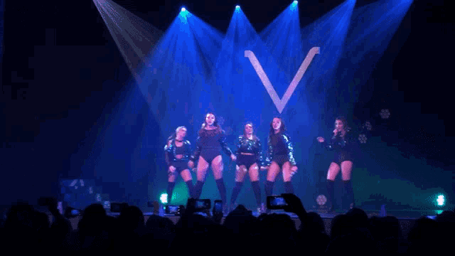 a group of women are dancing on a stage in front of a blue v