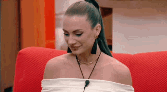 a woman is sitting on a red couch wearing a white off the shoulder top and earrings .