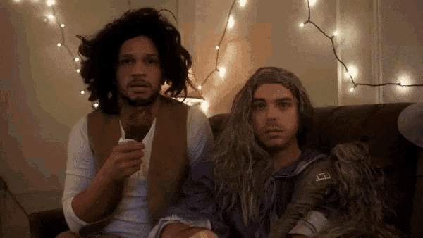 two men sitting on a couch one of whom is wearing a wig