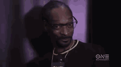snoop dogg is wearing glasses and a black shirt while talking into a microphone in a dark room .