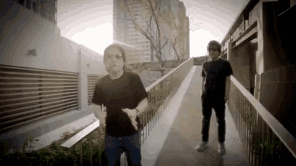 two men standing on a walkway with the sun shining through the buildings behind them