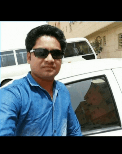 a man wearing sunglasses and a blue shirt stands in front of a white car