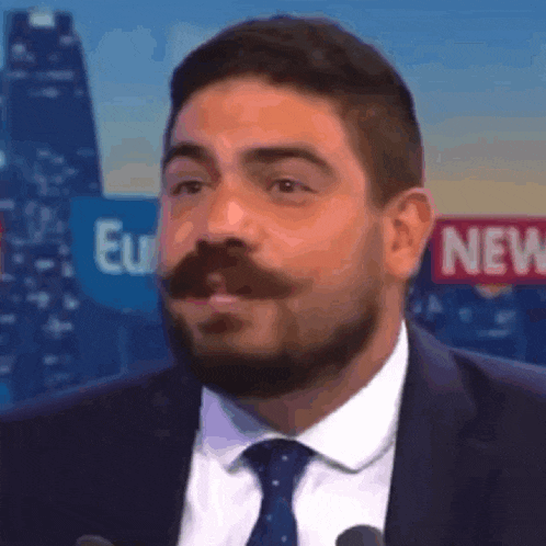 a man with a beard in a suit and tie is standing in front of a sign that says new .