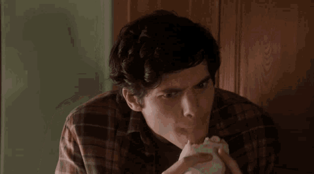 a young man in a plaid shirt is eating a sandwich
