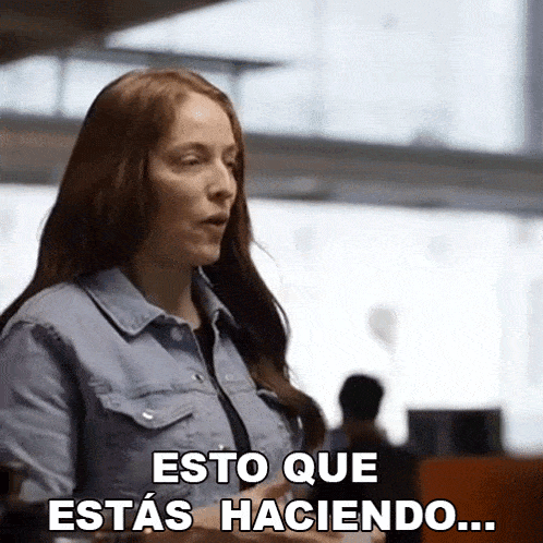 a woman in a denim shirt is standing in front of a window and talking in spanish .