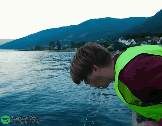 a person in a yellow vest is drinking water from a lake with mountains in the background
