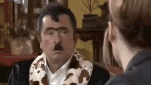 a man with a mustache is talking to a woman while wearing a cow print scarf around his neck .
