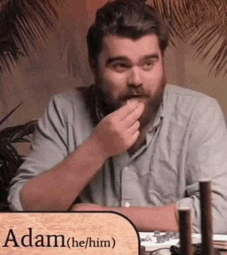 a man with a beard is sitting at a table with a sign that says adam he him