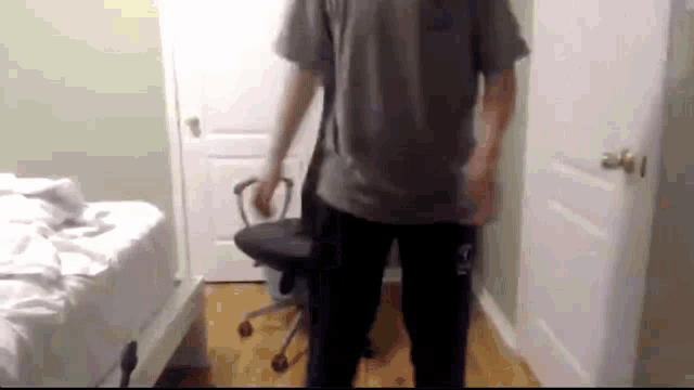 a man in a grey shirt is standing next to an office chair in a room .