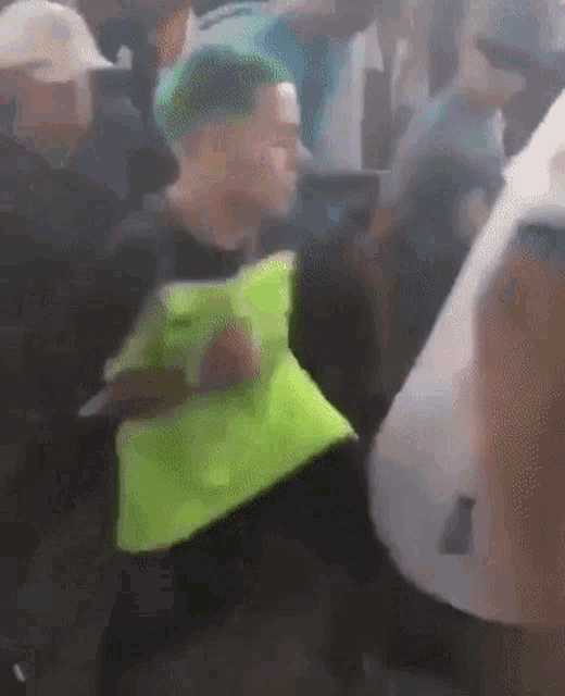 a man with green hair is standing in a crowd of people wearing a yellow vest .